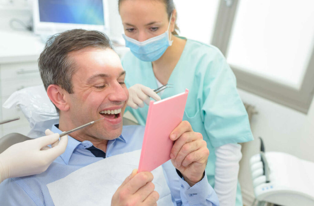 A man in a light blue long-sleeve collared shirt is holding a pink rectangular mirror and his mouth open while checking his newly installed dental bridges. The man is sitting on a dental chair while the female dentist is sitting on a stool on the side.