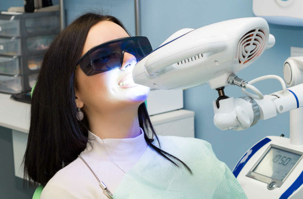 A young woman is sitting on a dental chair while her teeth are getting whitened with a use of UV light.