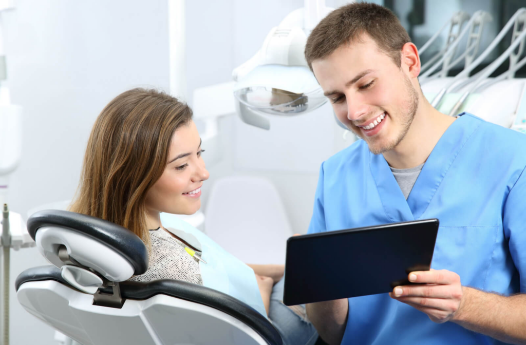 A male dentist in blue scrubs and a woman sitting in a dentist's chair looking at a tablet and discussing Invisalign.