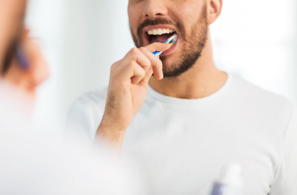 A man wearing white shirt with a toothbrush in his mouth smiling as he cleans his teeth.
