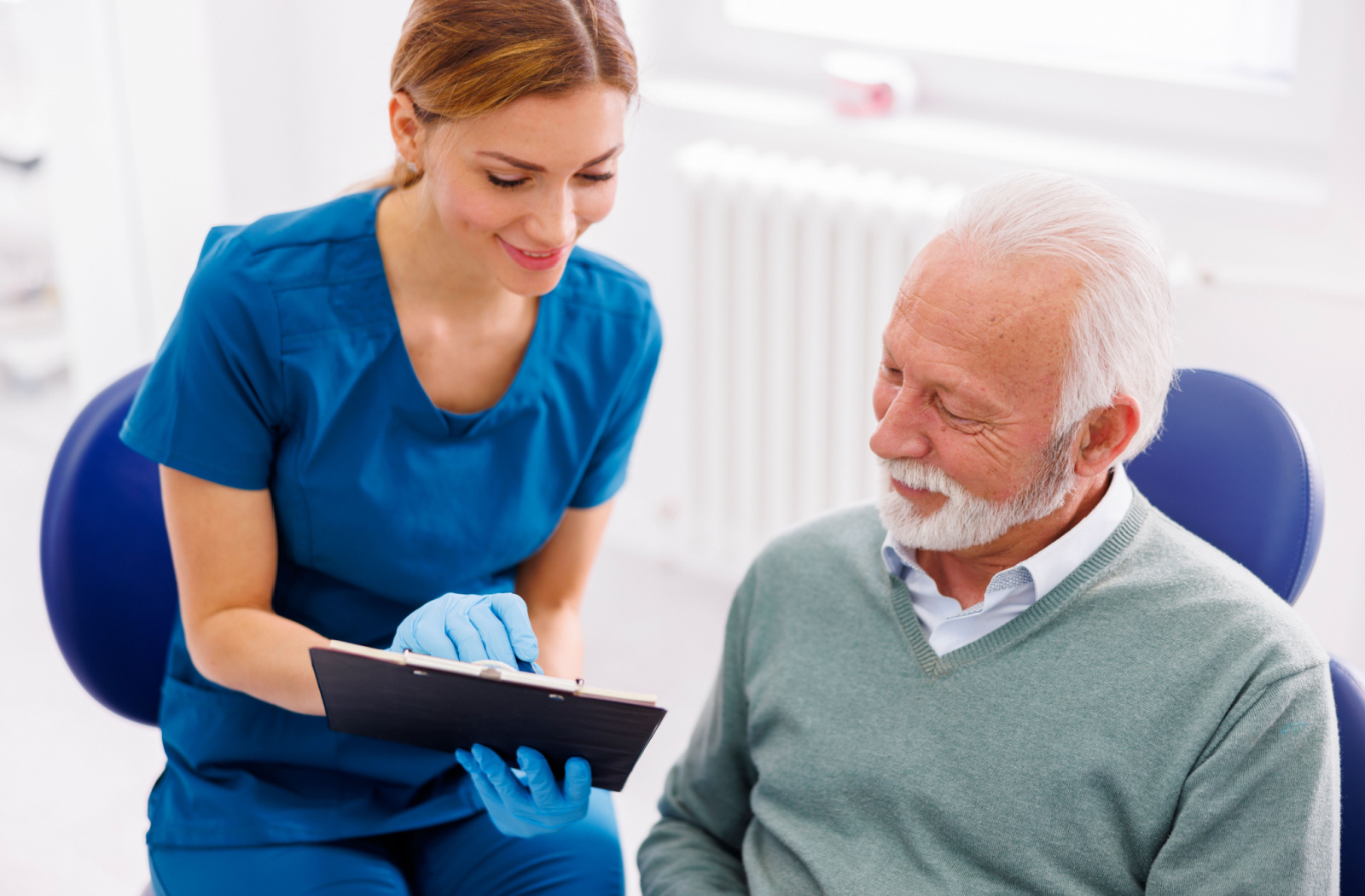 A female dentist in blue scrubs holding a clipboard and showing it to a senior man.