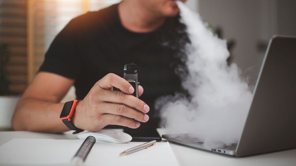 A man holds an e-cigarette and exhales vapour while working at his laptop