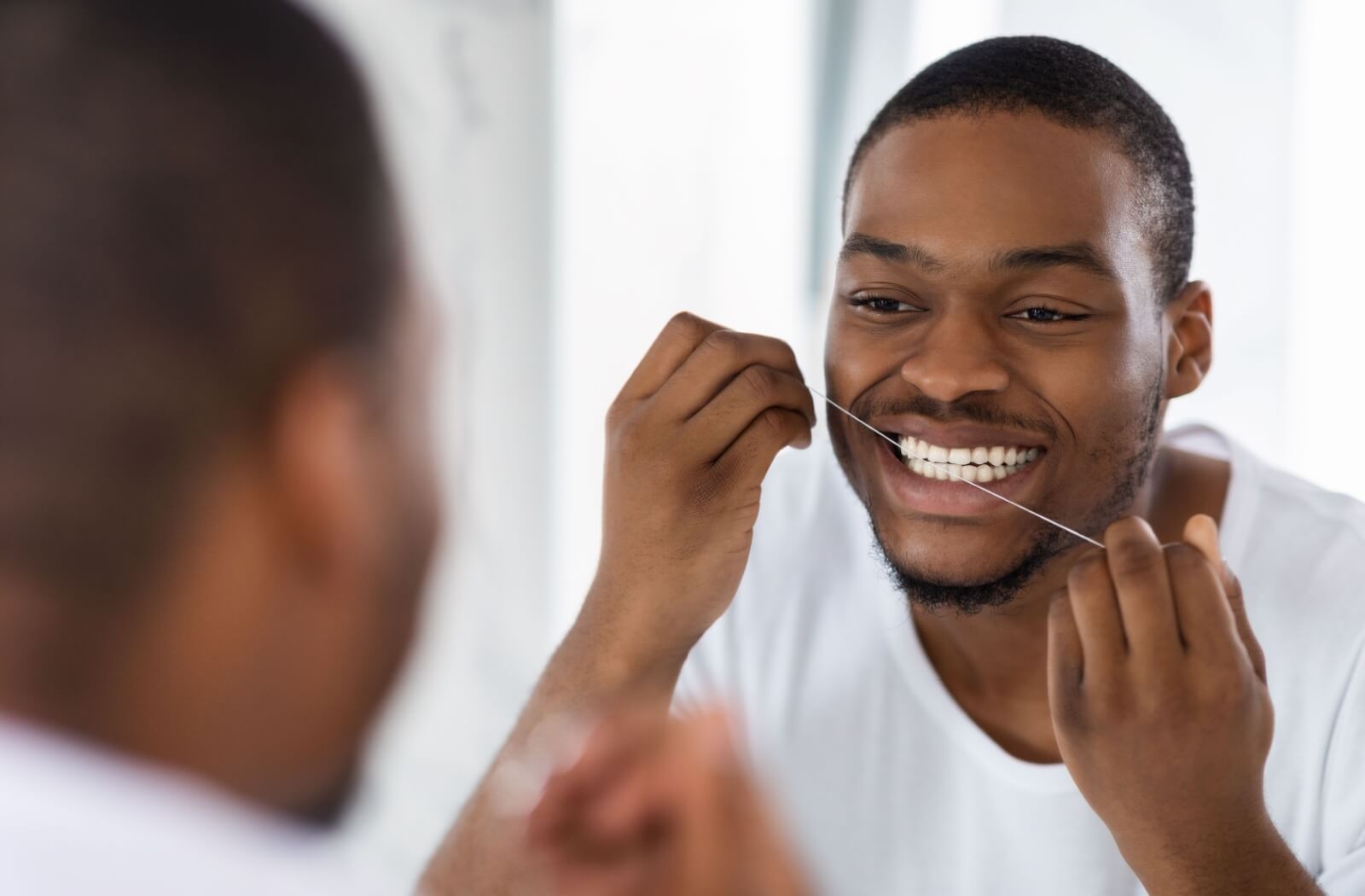 A man in a white shirt flossing his teeth in front of a mirror.