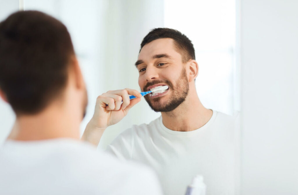 A man in a white shirt brushing his teeth in front of his bathroom mirror.