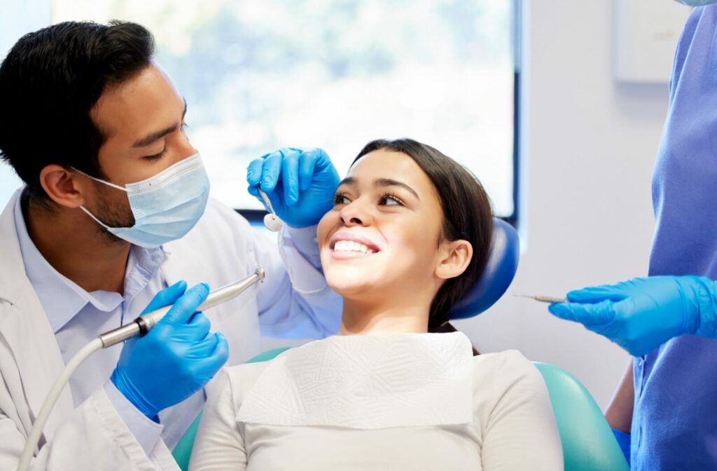 A smiling young woman sits in the dentist chair undergoing an exam with her dentist.