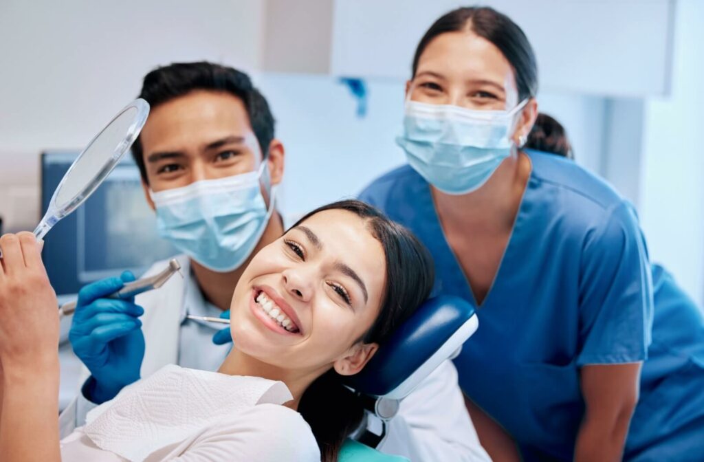 A patient lays in a dental chair, holds a mirror, and smiles. Behind her, two dental professionals smile behind dental masks.