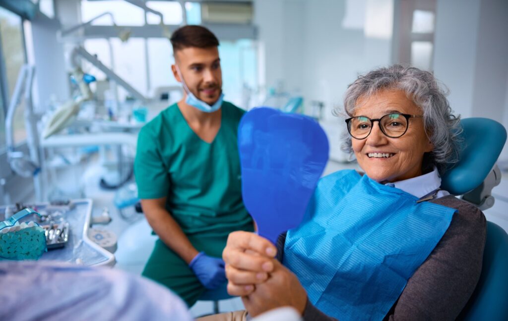 A senior sitting in a dentist's offices smiles and admires their brand new dentures in a hand mirror.