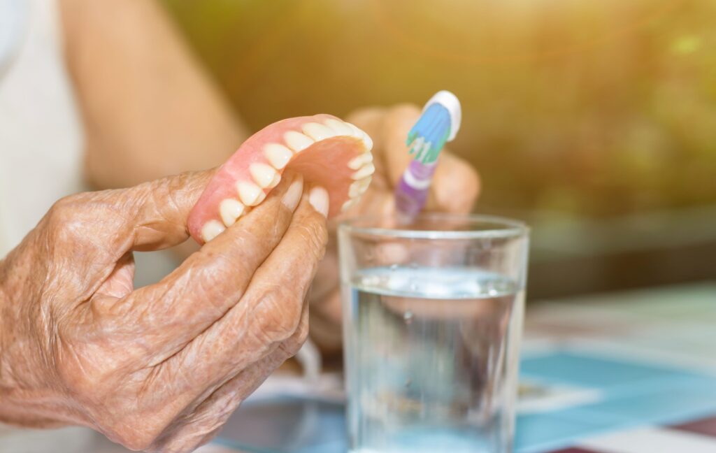 A person uses a toothbrush to gently clean their dentures before placing them in a glass of clean water.