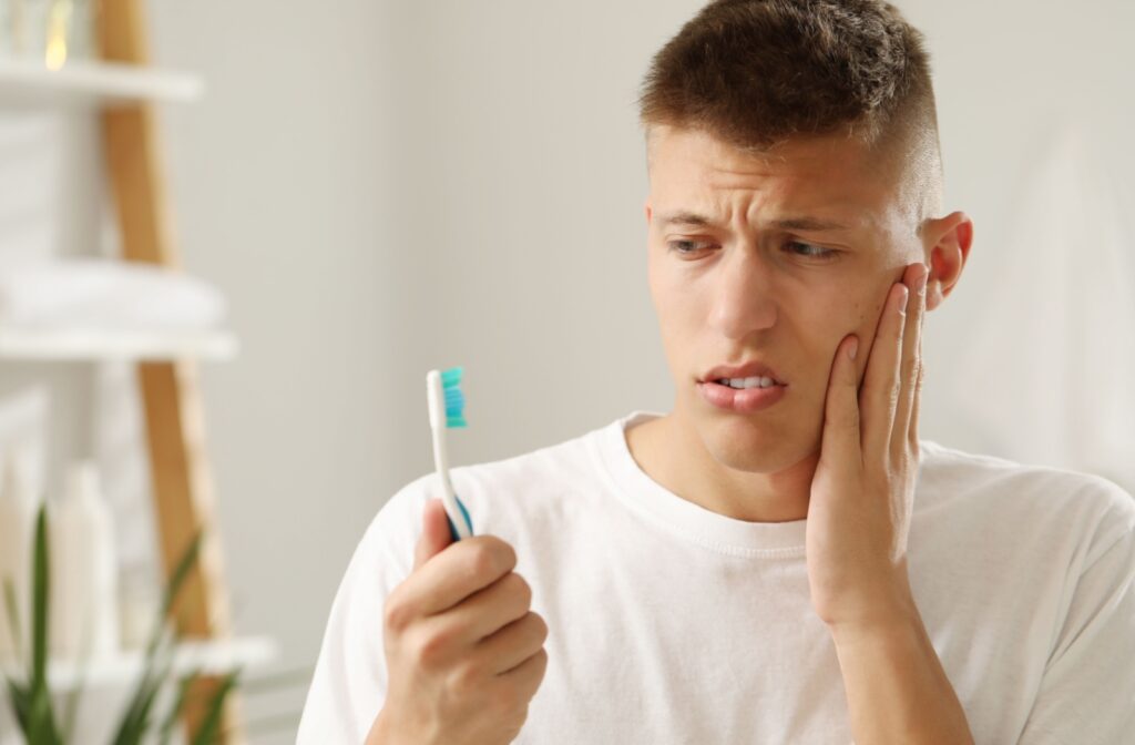 A young adult holds their cheek while looking at their toothbrush, worried about disturbing their tooth extraction recovery.