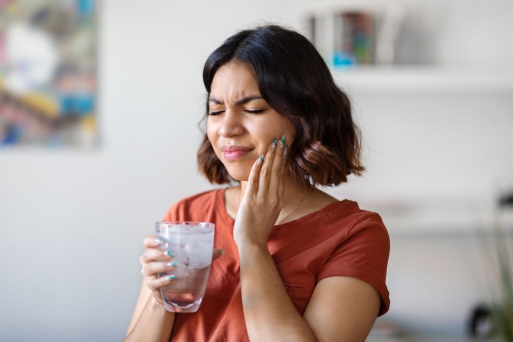 Woman holding a glass of ice water and touching her cheek due to tooth sensitivity.